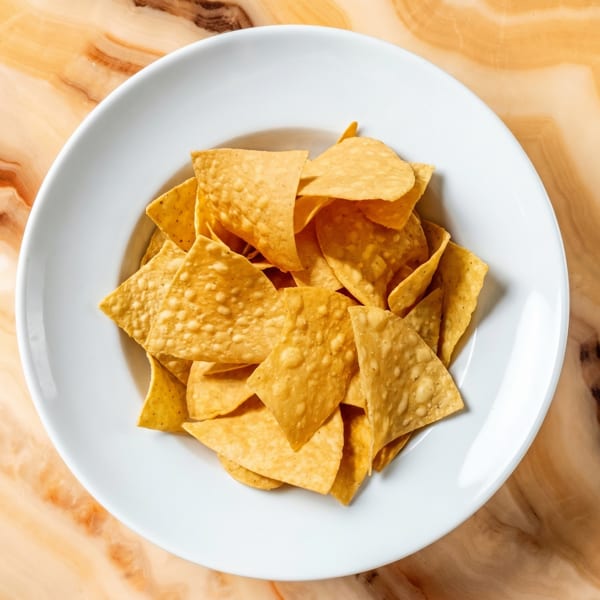 A white bowl filled with golden tortilla chips sits on a polished, light brown marble surface.