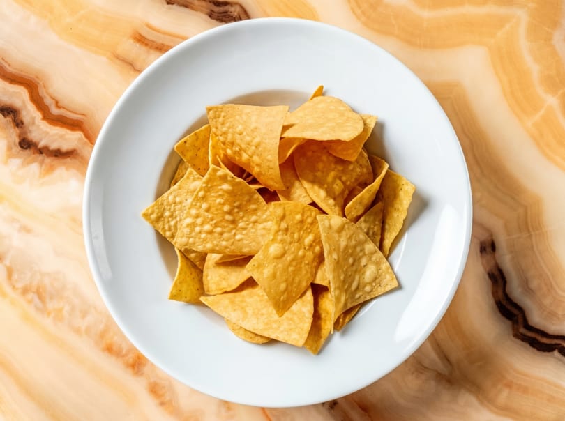 A white bowl filled with golden tortilla chips sits on a polished, light brown marble surface.