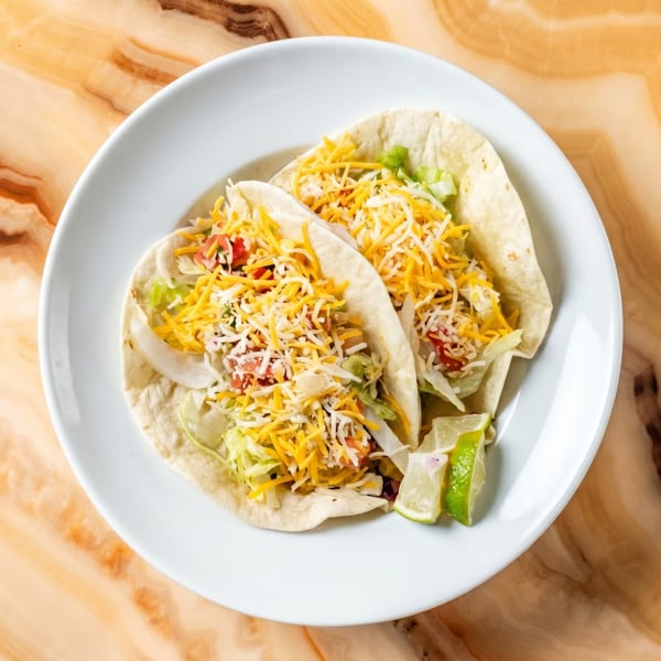 Two soft tacos with shredded cheese, lettuce, and tomatoes, served with lime wedges on a white plate.
