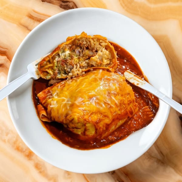 A beef and rice burrito, cut in half, covered in red sauce and melted cheese on a white plate.