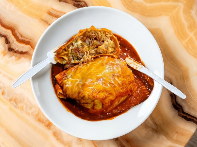 A beef and rice burrito, cut in half, covered in red sauce and melted cheese on a white plate.