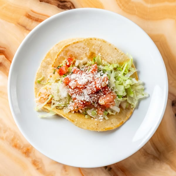 A single taco on a white plate, topped with lettuce, diced tomatoes, and cotija cheese.